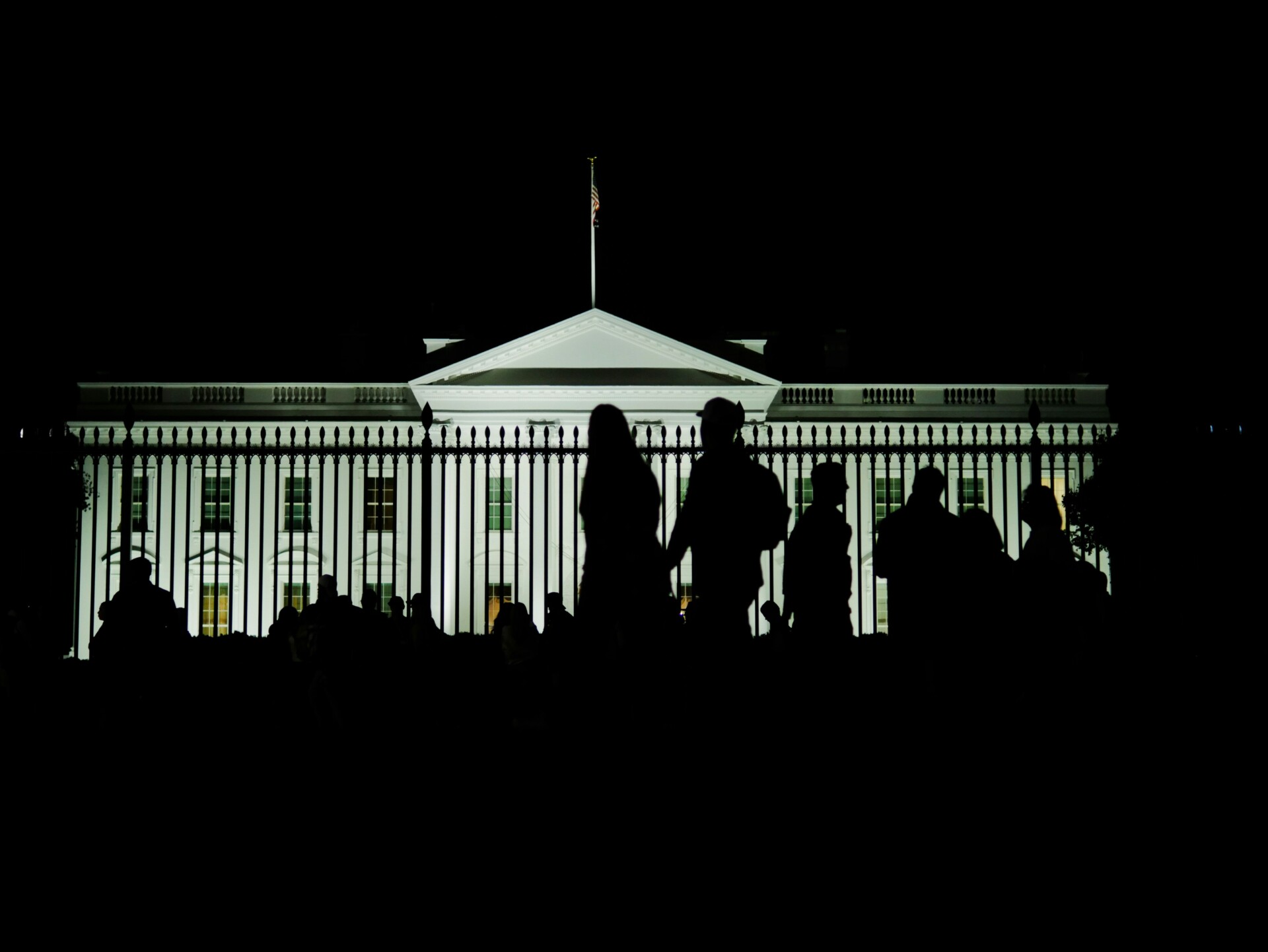 Image of the White House in Washington, DC at night, with pedestrians walking across the frame